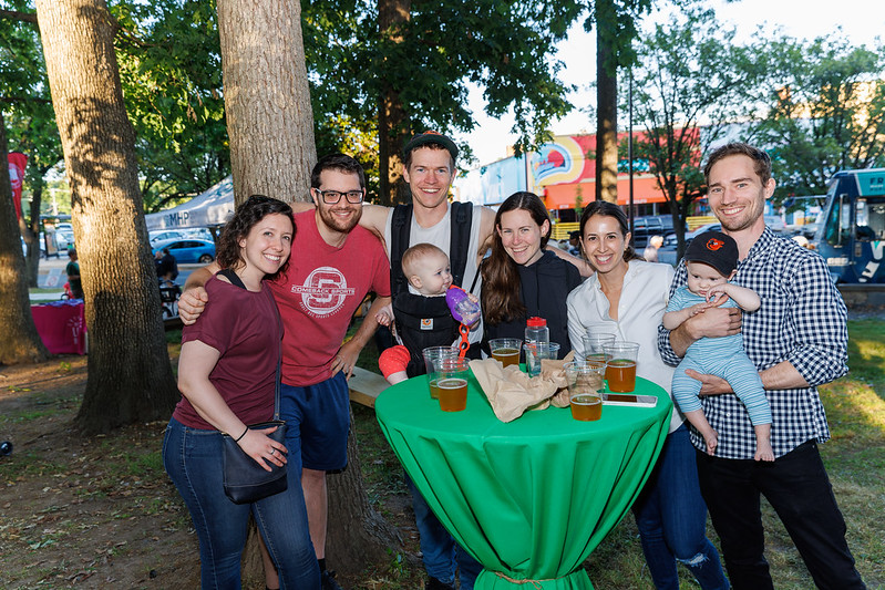 Smiling adults with babies standing around table with beers